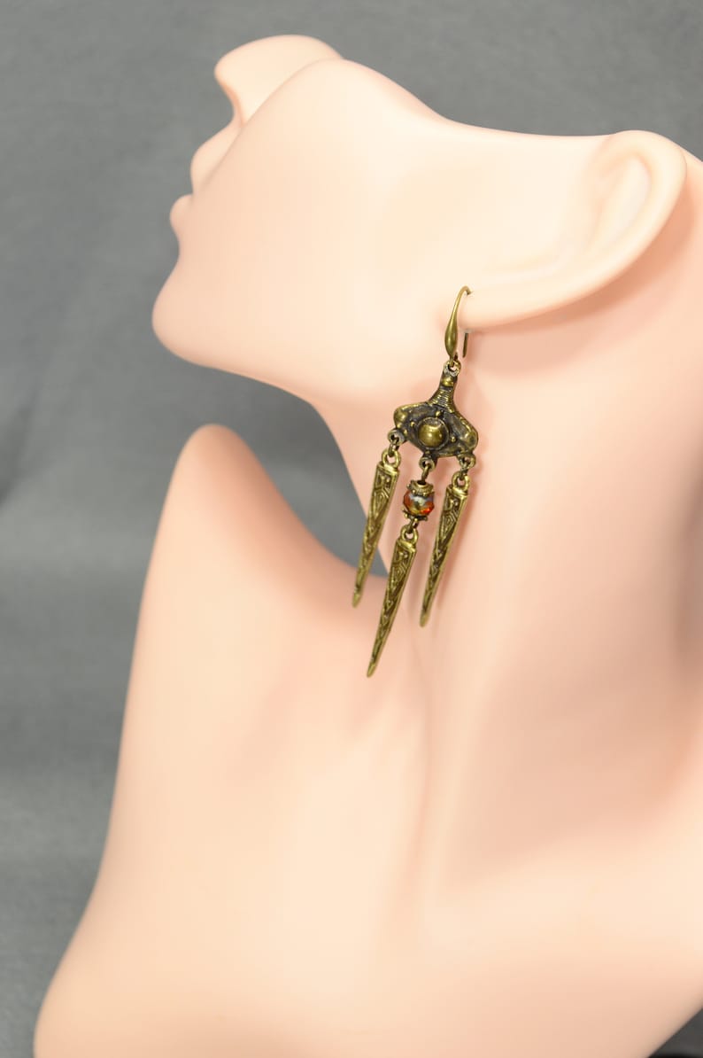 May include: Gold tone dangle earrings with a geometric design and a single red bead. The earrings are on a mannequin's ear.