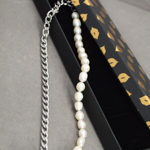 May include: A two-tone necklace featuring a silver chain on one side and a strand of white pearls on the other. The necklace is displayed in a black box with a gold lip pattern.