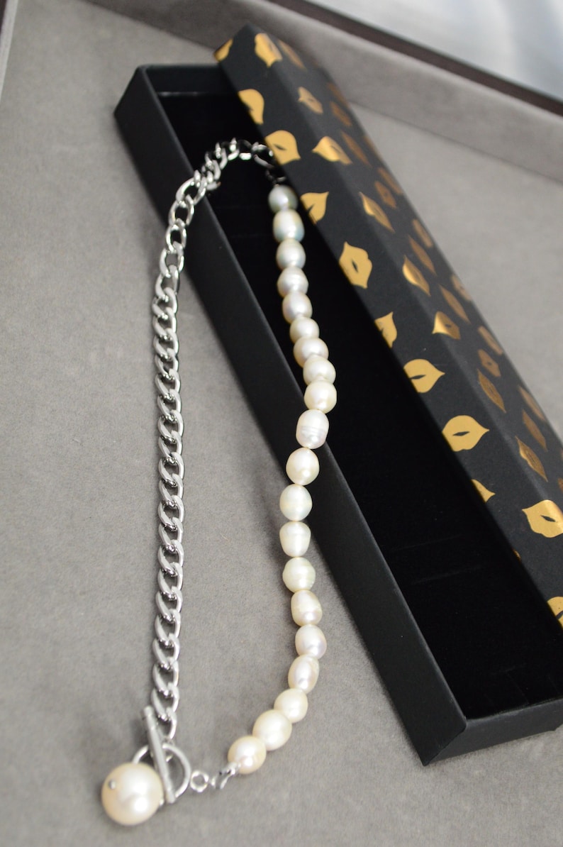May include: A two-tone necklace featuring a silver chain on one side and a strand of white pearls on the other. The necklace is displayed in a black box with a gold lip pattern.