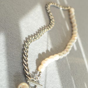 May include: A necklace featuring a combination of a silver chain and a strand of pearls. The necklace has a toggle clasp and a single pearl pendant. The pearls are white and the chain is silver.