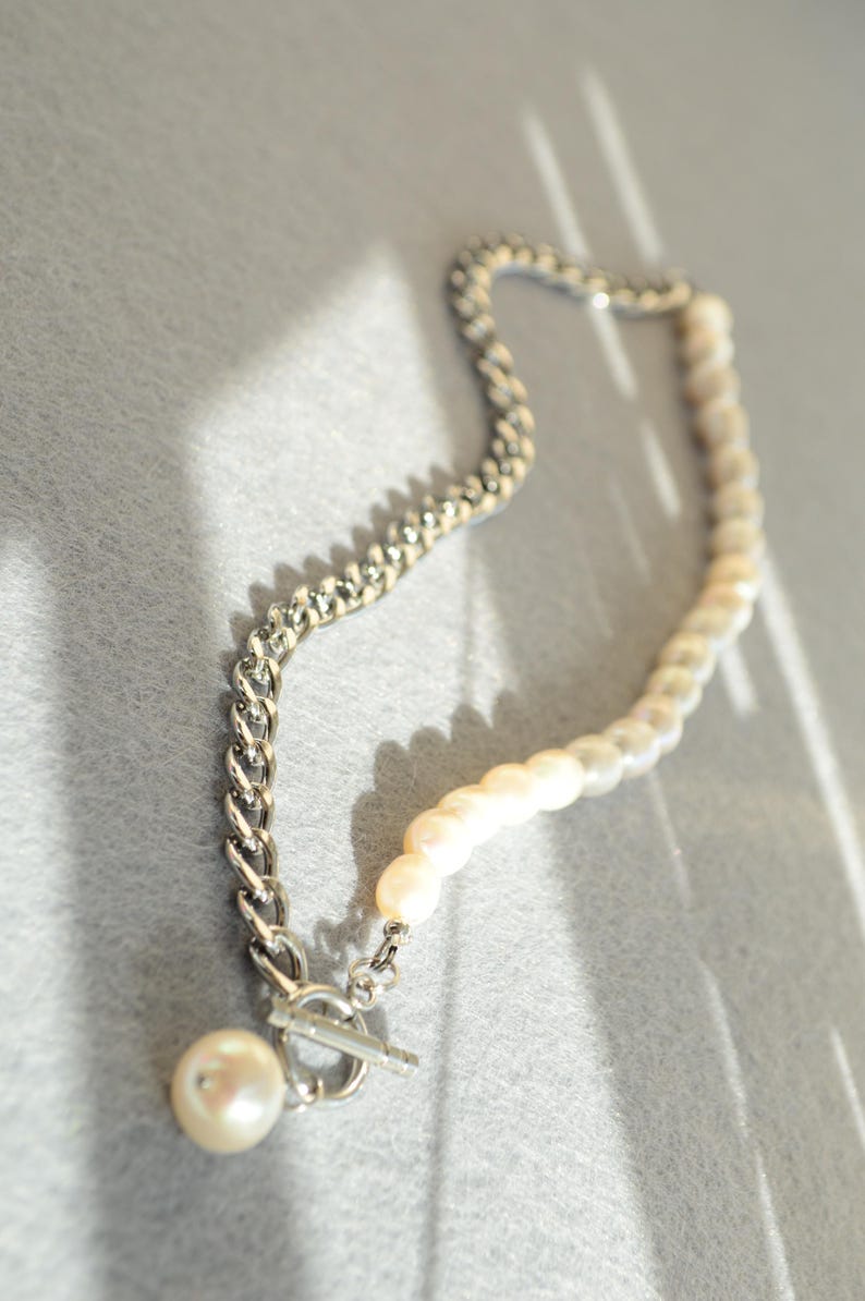 May include: A necklace featuring a combination of a silver chain and a strand of pearls. The necklace has a toggle clasp and a single pearl pendant. The pearls are white and the chain is silver.