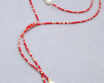 May include: A lariat-style necklace featuring a long strand of small red, white, and gold-toned beads. The necklace includes a single round white pearl accent on the upper loop and two elongated white baroque pearls at the ends of the drop.