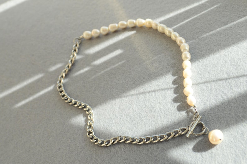 May include: A necklace featuring a combination of pearls and a silver chain. The necklace has a toggle clasp and a single pearl pendant. The pearls are off-white, and the chain is a silver color.