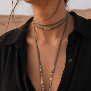 Peut inclure: Un collier ras du cou en daim gris à trois rangs. Les deux rangs supérieurs comportent de petites perles argentées. Le rang inférieur comporte deux pendentifs argentés avec des gravures décoratives. La personne porte une chemise noire boutonnée.