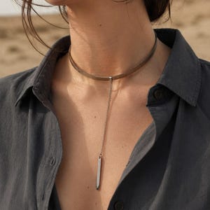 May include: A metallic choker necklace featuring a thin, flat band that sits close to the neck. A delicate, slender chain drops from the center of the band, ending in a small, polished rectangular bar pendant that hangs vertically against the chest.