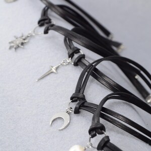 May include: Black leather chokers with silver-tone charms. The charms include a crescent moon, a sunburst, a cross, and a pearl. The chokers are knotted and have a minimalist design, suitable for everyday wear.