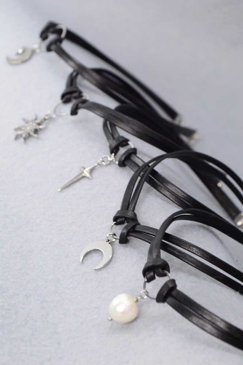 May include: Black leather chokers with silver-tone charms. The charms include a crescent moon, a sunburst, a cross, and a pearl. The chokers are knotted and have a minimalist design, suitable for everyday wear.