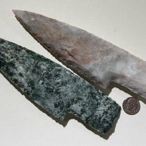 May include: Two stone arrowheads, one green and one tan, with a coin for scale. The green arrowhead has a textured surface. The tan arrowhead has red and white marbling. Both have a notched base.