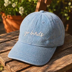 Peut inclure: Une casquette de baseball en jean bleu clair avec une visière incurvée et le texte en cursive blanche brodé sur le devant qui indique go birds. La casquette présente une construction à six panneaux avec des œillets cousus et un bouton au sommet.