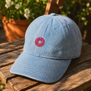 Puede incluir: Una gorra de béisbol de mezclilla azul claro con un dónut rosa bordado y chispas de colores en el panel frontal. La gorra cuenta con una visera curva, un botón en la parte superior y ojales cosidos para la ventilación en un diseño clásico.