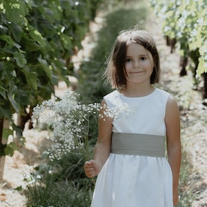 Può includere: Una bambina con un vestito bianco e una cintura grigia tiene un mazzo di fiori bianchi mentre è in piedi in un vigneto.
