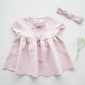 May include: A pale pink linen dress with short sleeves and a tie at the neckline. The dress has a gathered skirt and is perfect for a special occasion.