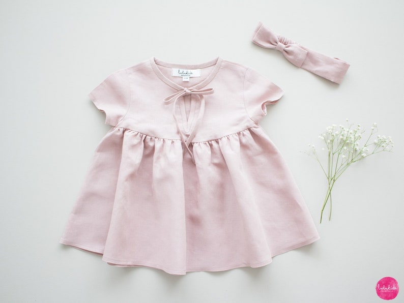 May include: A pale pink linen dress with short sleeves and a tie at the neckline. The dress has a gathered skirt and is perfect for a special occasion.