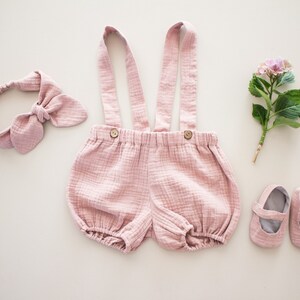 以下が含まれることがあります： ピンクのベビー服。ヘッドバンド、ブルマ、サスペンダー付き。柔らかく軽い生地でできており、女の子の赤ちゃんに最適です。