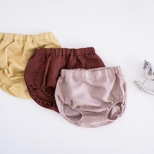 May include: Three pairs of baby bloomers in different colors: mustard yellow, burgundy, and light pink. The bloomers are made of a soft, lightweight fabric and have a gathered elastic waistband.
