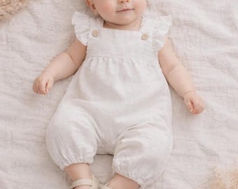 May include: A baby wearing a white linen romper with ruffle sleeves and wooden button details. The baby is also wearing cream-colored Mary Jane shoes. The romper is a soft, neutral color, perfect for a baby's wardrobe.