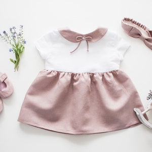 May include: A white and blush pink linen dress with a peter pan collar and a bow at the neckline. The dress has short sleeves and a gathered skirt. The dress is paired with a blush pink headband and a pair of white shoes.