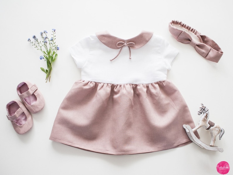 May include: A white and blush pink linen dress with a peter pan collar and a bow at the neckline. The dress has short sleeves and a gathered skirt. The dress is paired with a blush pink headband and a pair of white shoes.