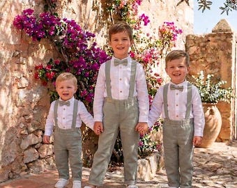 Op de afbeelding: Drie jonge jongens in bijpassende outfits. Elk draagt een wit overhemd, een vlinderdas, bretels en een lichtgroene broek. Ze staan voor een stenen muur versierd met felroze bloemen.