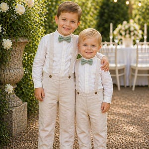 May include: Two young boys in matching outfits. They wear white shirts, beige trousers with braces, and sage green bow ties. They are outside, smiling, in front of a hedge. The trousers are a light beige colour.