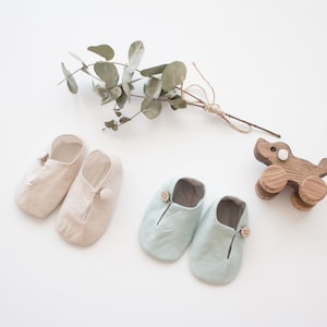 Op de afbeelding: Twee paar babyschoentjes, één paar is beige en het andere is lichtblauw. Beide paren hebben een knoopdetail. Er is ook een houten speelgoedhondje op de foto.