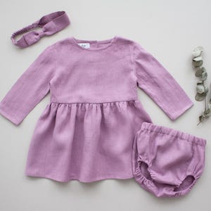 May include: A lavender linen dress with long sleeves and a gathered skirt, paired with matching lavender linen bloomers and a lavender headband.