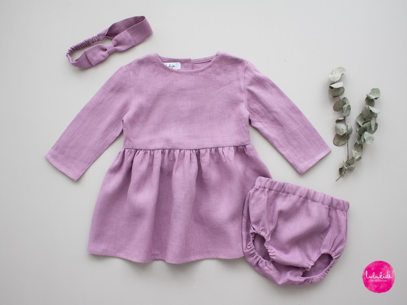 May include: A lavender linen dress with long sleeves and a gathered skirt, paired with matching lavender linen bloomers and a lavender headband.
