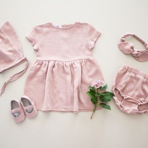以下が含まれることがあります： ピンクのベビー服セットには、ドレス、ヘッドバンド、ボンネット、ブルマ、靴が含まれています。ドレスは半袖で、スカートはギャザーが入っています。ヘッドバンドはリボンで結ばれています。ボンネットは後ろにリボンが付いています。ブルマはウエストがギャザーで、足首はゴムになっています。靴はメリージェーンスタイルです。
