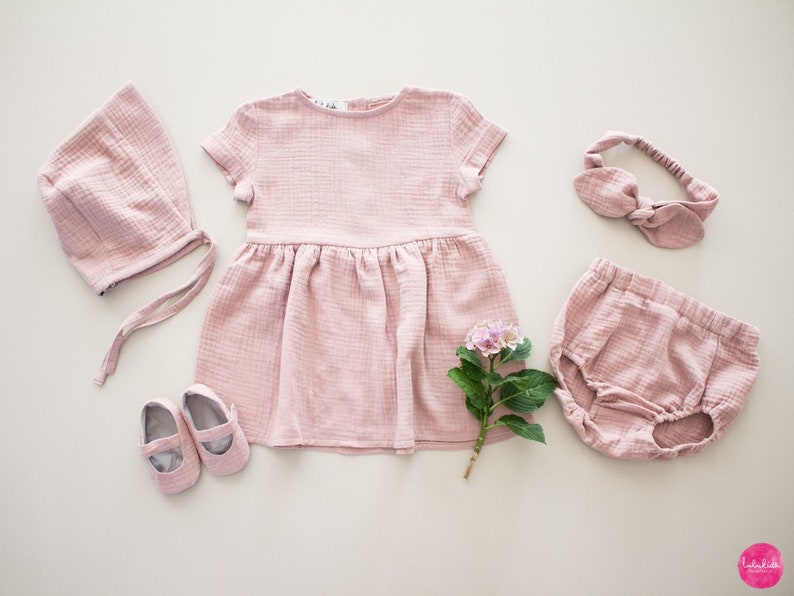 以下が含まれることがあります： ピンクのベビー服セットには、ドレス、ヘッドバンド、ボンネット、ブルマ、靴が含まれています。ドレスは半袖で、スカートはギャザーが入っています。ヘッドバンドはリボンで結ばれています。ボンネットは後ろにリボンが付いています。ブルマはウエストがギャザーで、足首はゴムになっています。靴はメリージェーンスタイルです。