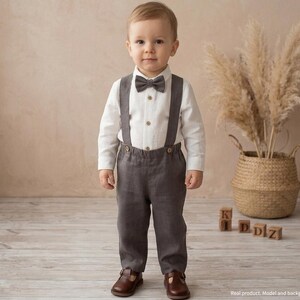 May include: A young child in a formal outfit, including a white button-down shirt, gray bow tie, suspenders, and matching gray pants. The child is wearing brown shoes. The outfit is suitable for a special occasion. The background includes a decorative plant and wooden blocks.