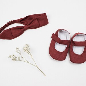 May include: A set of baby shoes and a headband in a burgundy color. The shoes are made of a soft fabric and have a simple design. The headband is a simple bow style.