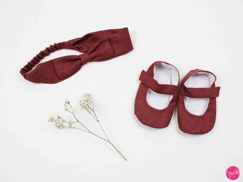 May include: A set of baby shoes and a headband in a burgundy color. The shoes are made of a soft fabric and have a simple design. The headband is a simple bow style.