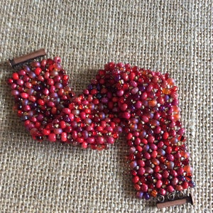 Op de afbeelding: Handgemaakte armband bestaande uit kleine, levendige rode kralen. De kralen zijn gerangschikt in een geweven patroon, waardoor een textuur ontstaat. De armband heeft een koperkleurige sluiting. Het ontwerp is rijk aan kleur en detail.