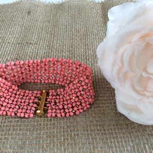 May include: A pink beaded bracelet with a gold clasp. The bracelet is made of small, round beads and is shown on a hessian background.