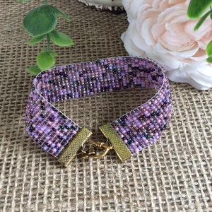 Puede incluir: Un brazalete de cuentas de color morado y rosa con un cierre dorado. El brazalete está hecho de pequeñas cuentas de colores y tiene un diseño simple y elegante.