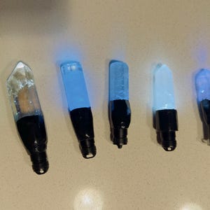 May include: Seven illuminated crystal-like pendants arranged in a row. Each piece features a clear or translucent pointed top attached to a black cylindrical base. The crystals emit various shades of blue, white, and green light from within.