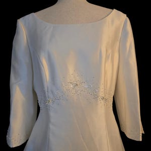 May include: Ivory-colored wedding dress with a boat neckline and three-quarter sleeves. The waistline is embellished with delicate beading and sequins. The dress is displayed on a mannequin against a black background.