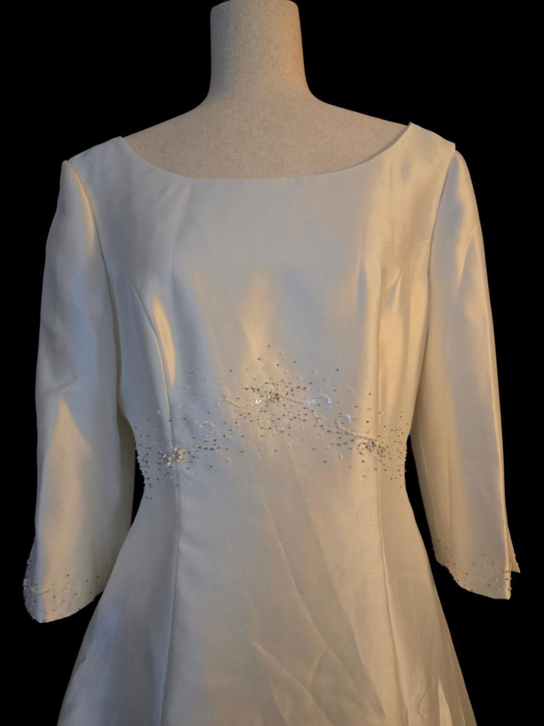 May include: Ivory-colored wedding dress with a boat neckline and three-quarter sleeves. The waistline is embellished with delicate beading and sequins. The dress is displayed on a mannequin against a black background.