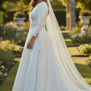 May include: A white wedding dress with long sleeves and a flowing skirt. The dress features a beaded belt and a long, sheer veil. The bride is standing in a garden setting with greenery and flowers in the background.
