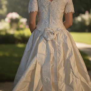 May include: An off-the-shoulder cream-coloured gown featuring short sleeves, intricate lace embroidery on the bodice, a central back zip, and a large fabric rose at the waist with cascading vertical panels adorned with small floral appliqu&eacute;s.