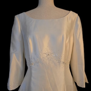 May include: Ivory-colored wedding dress with a boat neckline and three-quarter sleeves. The waistline is embellished with a delicate beaded design. The dress is displayed on a mannequin against a black background.