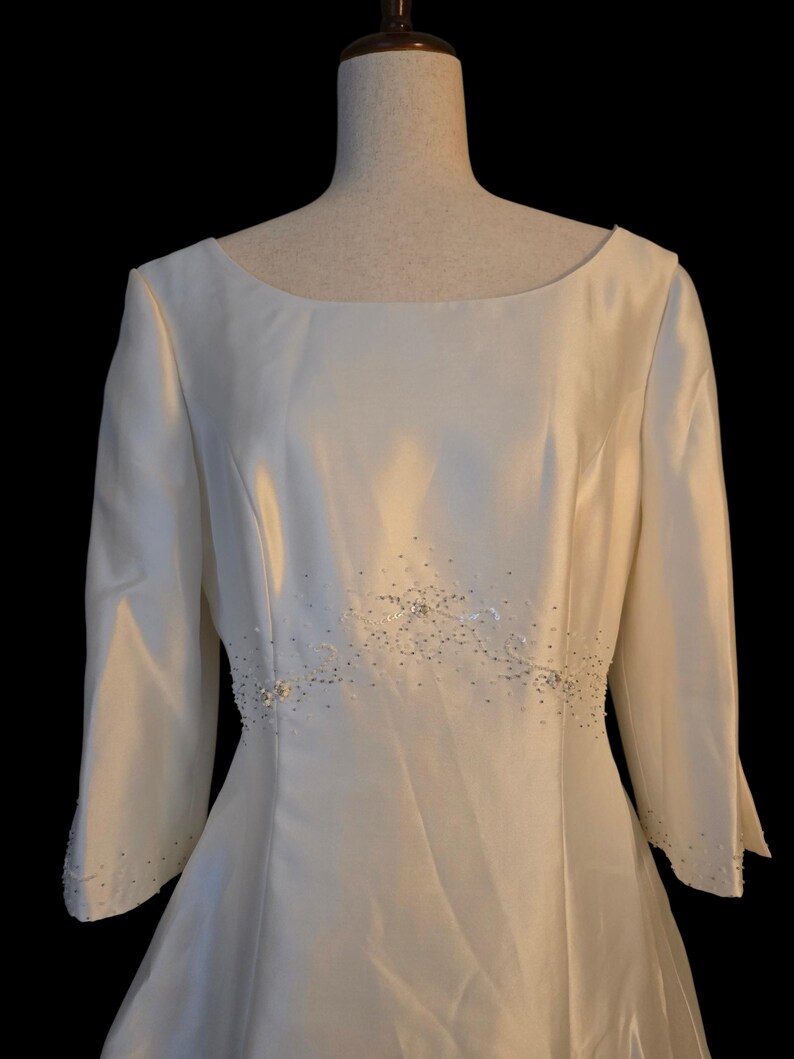 May include: Ivory-colored wedding dress with a boat neckline and three-quarter sleeves. The waistline is embellished with a delicate beaded design. The dress is displayed on a mannequin against a black background.
