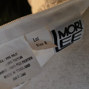 May include: A white garment label displaying the brand name Mori Lee, size 8, and fabric composition. The text specifies 70% silk and 30% rayon for the shell, 100% polyester for the lining, and states that the garment was made in Thailand.
