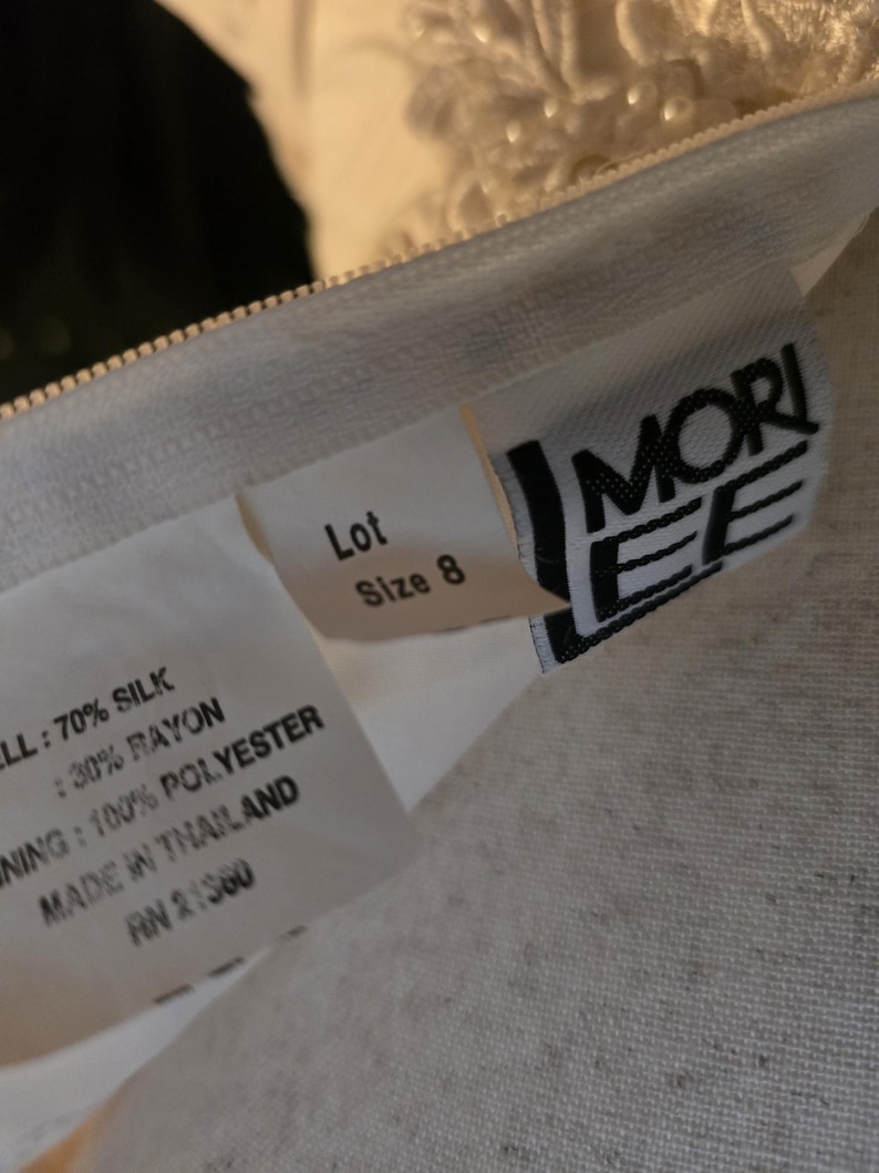 May include: A white garment label displaying the brand name Mori Lee, size 8, and fabric composition. The text specifies 70% silk and 30% rayon for the shell, 100% polyester for the lining, and states that the garment was made in Thailand.