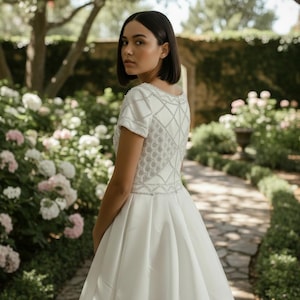 May include: A white wedding dress with a short-sleeved bodice featuring a geometric silver beaded design. The dress has a full skirt and is worn outdoors in a garden setting with a stone pathway and lush greenery.