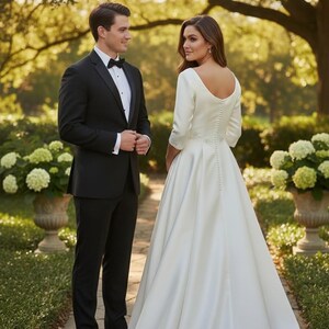 May include: A formal wedding scene featuring a couple on a stone path. The person on the left is wearing a black tuxedo with a bow tie. The person on the right is wearing a white satin wedding gown with a long train and long sleeves. The background includes greenery and large planters.