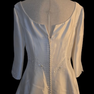 May include: Ivory-colored bridal gown with a scoop neckline and three-quarter sleeves. The back features a row of buttons and subtle beaded embellishments. The dress is displayed on a mannequin against a black background.