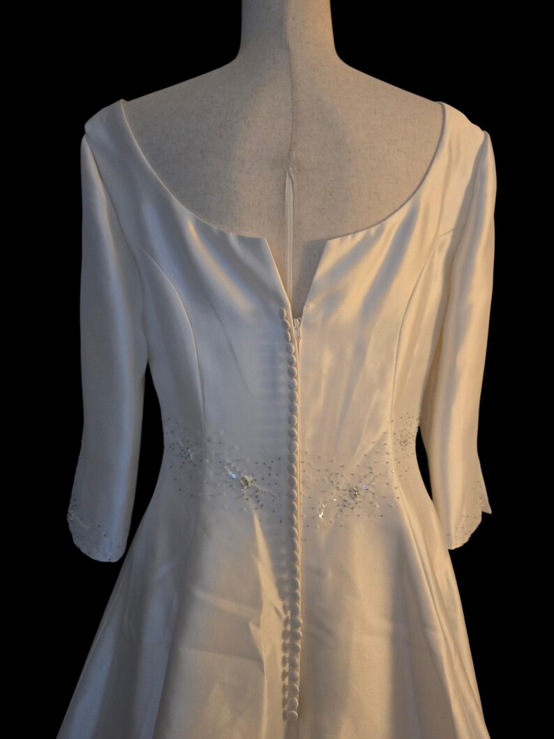 May include: Ivory-colored bridal gown with a scoop neckline and three-quarter sleeves. The back features a row of buttons and subtle beaded embellishments. The dress is displayed on a mannequin against a black background.