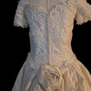 May include: An off-the-shoulder cream-coloured gown featuring an intricate bodice with white floral lace appliques, sequins, and small pearls. The waist is accented with a large fabric rose and smaller floral details, leading into a full skirt.