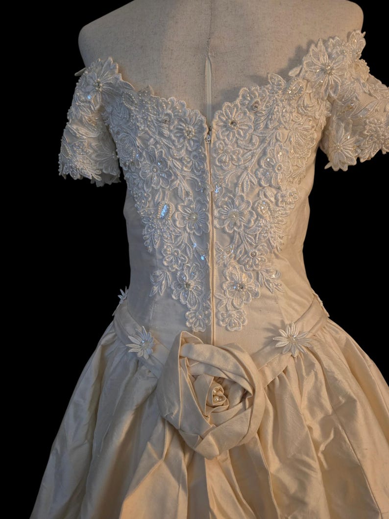 May include: An off-the-shoulder cream-coloured gown featuring an intricate bodice with white floral lace appliques, sequins, and small pearls. The waist is accented with a large fabric rose and smaller floral details, leading into a full skirt.